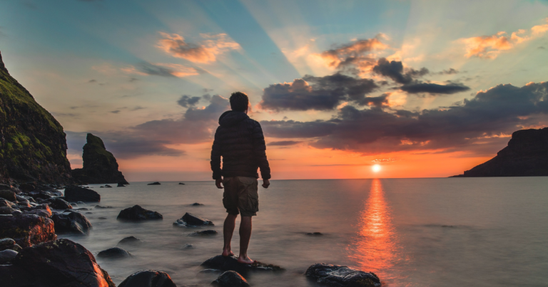 Older, likely retired, man watching the sunet by the ocean standing on rocks